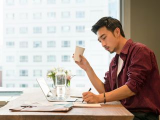 An asian man in front of his laptop writing in a notebook