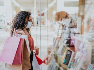 A woman with a face mask on holding shopping bags window shopping