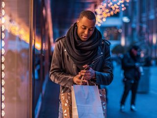 Man wearing winter clothes standing on a street holding paper bags and using a smartphone