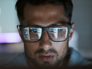 A focused man wearing glasses with computer code reflecting in the lenses.