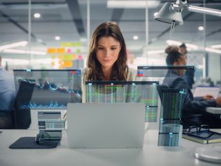 A woman working in an office surrounded by multiple digital screens displaying financial data and charts.