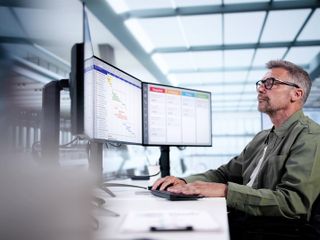 An individual seated at a desk, engaging with two computer monitors displaying project management data and charts in a modern office environment.