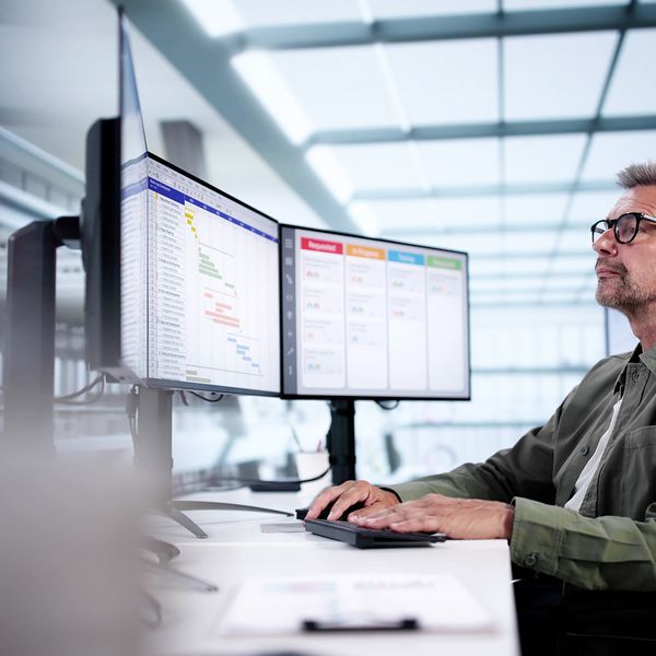 An individual seated at a desk, engaging with two computer monitors displaying project management data and charts in a modern office environment.