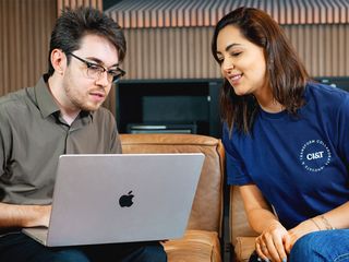 A man and a woman in a casual office setting look at a laptop screen together while sitting on a brown leather couch.