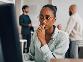 A young Black woman wearing glasses and a light green shirt looks thoughtfully at a computer screen in a bright office while colleagues converse in the background.